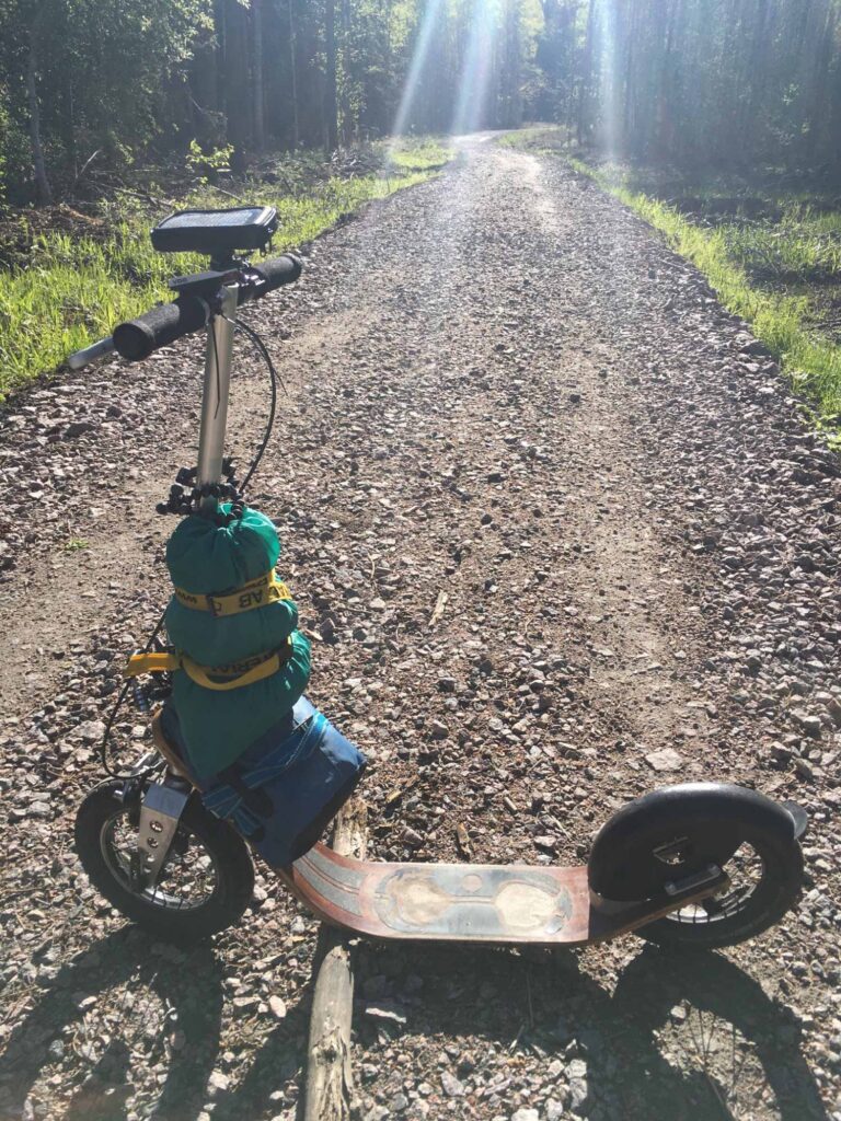 fully loaded boardy kick scooter with gear strapped, standing on a remote gravel road in sunlight.