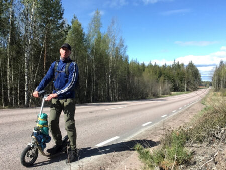 Man with a backpack standing beside a loaded Boardy kick scooter on a rural highway, surrounded by Nordic forest under a clear blue sky.