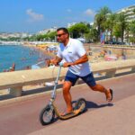 Man riding a Boardy kick scooter along a sunny seaside promenade.