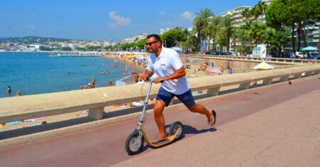 man riding a boardy kick scooter along a sunny seaside promenade.