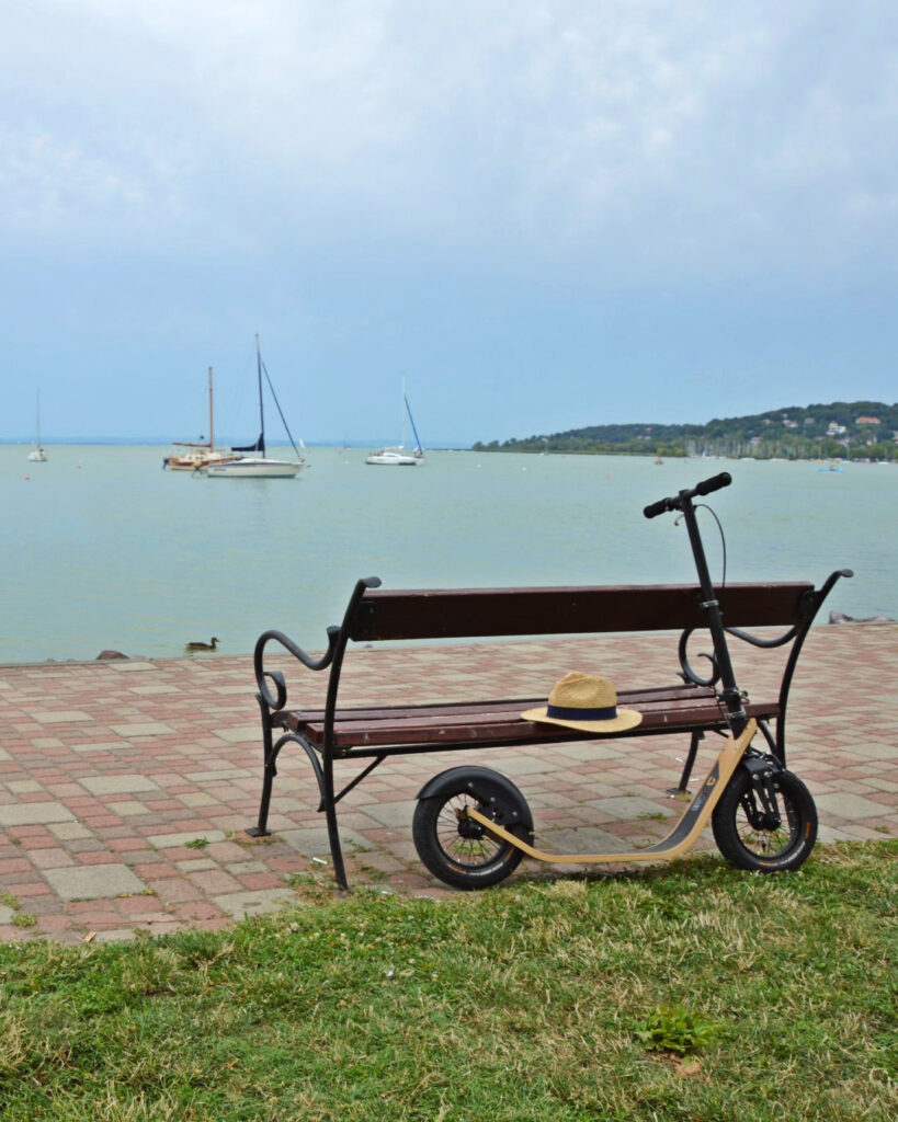 a parked boardy, a human powered scooter resting beside a wooden bench by the lakeshore, with sailboats anchoring the water of lake balaton in the background - boardscooter content hub