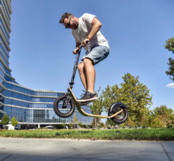 Strong Frame. Smart Flex. But No Stunts, Please. man doing a jump on a boardy scooter in front of a modern building.