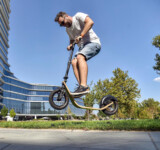 Strong Frame. Smart Flex. But No Stunts, Please. man doing a jump on a boardy scooter in front of a modern building.