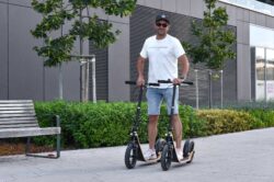 Because One Would Be Too Easy. man standing on two boardy scooters outdoors in a modern urban space.