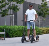 Because One Would Be Too Easy. man standing on two boardy scooters outdoors in a modern urban space.