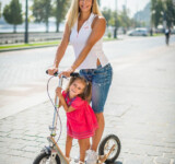 Room To Share woman riding a boardy scooter with a young child in front, both smiling.