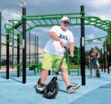 Go Big. Boardy Does. larger adult rider on a boardy scooter at a blue urban workout park.