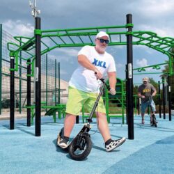 Go Big. Boardy Does. larger adult rider on a boardy scooter at a blue urban workout park.