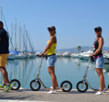 Crew On Deck three adults on wooden boardy kick scooters lined up by the marina, with yachts in the background.