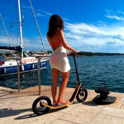 Barefoot. Beautiful. Boardy. woman in a beach cover-up standing on a boardy adult scooter by the seaside, with sailboats and calm blue water in the background on a sunny day.