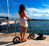 Barefoot. Beautiful. Boardy. woman in a beach cover-up standing on a boardy adult scooter by the seaside, with sailboats and calm blue water in the background on a sunny day.