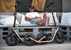 Parked Among Pallets man relaxing on a bench with two boardy adult scooters in front, showcasing their wide pneumatic tires and wooden decks.