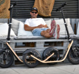 Parked Among Pallets man relaxing on a bench with two boardy adult scooters in front, showcasing their wide pneumatic tires and wooden decks.