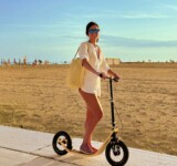 Where The Pavement Ends woman standing on a boardy kick scooter designed for adults, on a sunlit beach path.