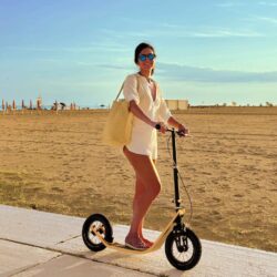 Where The Pavement Ends woman standing on a boardy kick scooter designed for adults, on a sunlit beach path.
