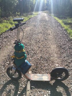 Rough Road Ahead fully loaded boardy kick scooter with gear strapped, standing on a remote gravel road in sunlight.
