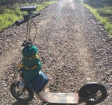 Rough Road Ahead fully loaded boardy kick scooter with gear strapped, standing on a remote gravel road in sunlight.