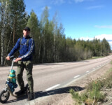 Endless Road. Trusted Ride. man with a backpack standing beside a loaded boardy kick scooter on a rural highway, surrounded by nordic forest under a clear blue sky.