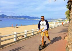 Baguette Run man riding a boardy kick scooter along a seaside promenade, holding a baguette and wearing a boardy logo shirt.
