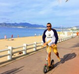 Baguette Run man riding a boardy kick scooter along a seaside promenade, holding a baguette and wearing a boardy logo shirt.