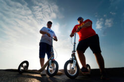 Boardy Bros two larger adult men riding boardy kick scooters, confidently posed against a bright sky.