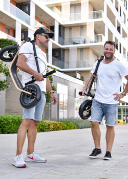 Two Boardys. No Rush. two men carrying folded boardy kick scooters over their shoulders in an urban environment.
