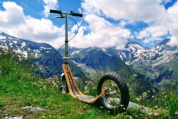 Go Beyond Pavement boardy all-terrain kick scooter resting on a grassy alpine slope with rugged mountain peaks and snow patches in the background.