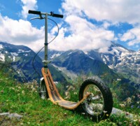 Go Beyond Pavement boardy all-terrain kick scooter resting on a grassy alpine slope with rugged mountain peaks and snow patches in the background.