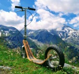Go Beyond Pavement boardy all-terrain kick scooter resting on a grassy alpine slope with rugged mountain peaks and snow patches in the background.