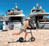 Fast Glide, Full Freedom. man riding a boardy kick scooter at high speed along a luxury marina with yachts in the background on a sunny day.