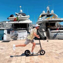 Fast Glide, Full Freedom. man riding a boardy kick scooter at high speed along a luxury marina with yachts in the background on a sunny day.