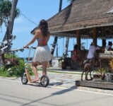 Tropical Ride Vibes woman riding a boardy scooter in a tropical resort area near bungalows.