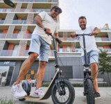 Neighborhood Ride two men riding their boardy scooters along a sunny residential street lined with apartment buildings.