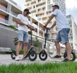 Street Chat two men standing face to face, chatting with their boardy scooters on a residential street lined with apartment buildings.
