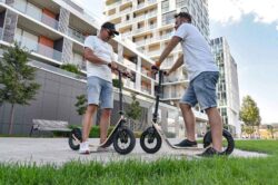 Street Chat two men standing face to face, chatting with their boardy scooters on a residential street lined with apartment buildings.