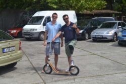 Wheelie Fun man with a prosthetic leg performing a wheelie on a boardy scooter in a parking lot.