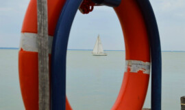 A red life ring hanging on a wooden post by tranquil Lake Balaton, with a sailboat drifting in the distance.