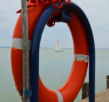 Life Buoy A red life ring hanging on a wooden post by tranquil Lake Balaton, with a sailboat drifting in the distance.