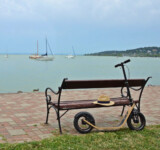 Rest Stop a parked boardy scooter resting beside a wooden bench by the lakeshore, with sailboats anchoring the water of lake balaton in the background.