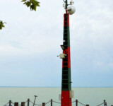 Beacon for Shores A weathered lighthouse structure beside Lake Balaton, red and green striped, with water stretching into the distance.