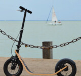 Scooter At The Shoreline a boardy kickscooter parked against a chain-link railing on the lakeside promenade, with sailboat visible over the water.